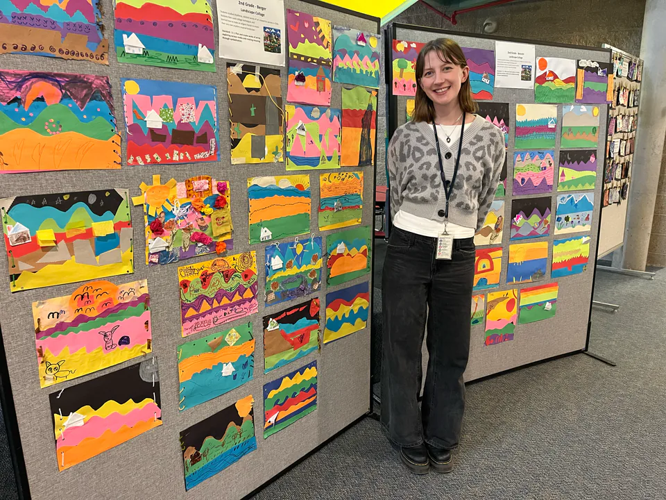 A teacher stands beside a corridor display board covered with children’s layered paper landscape collages.