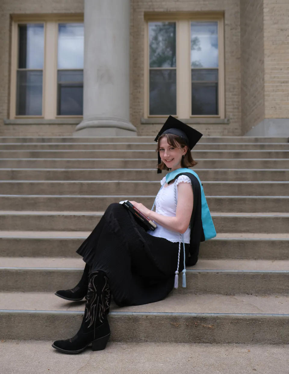 Sarah in a graduation cap and stole, sitting on the stone steps of a university building.