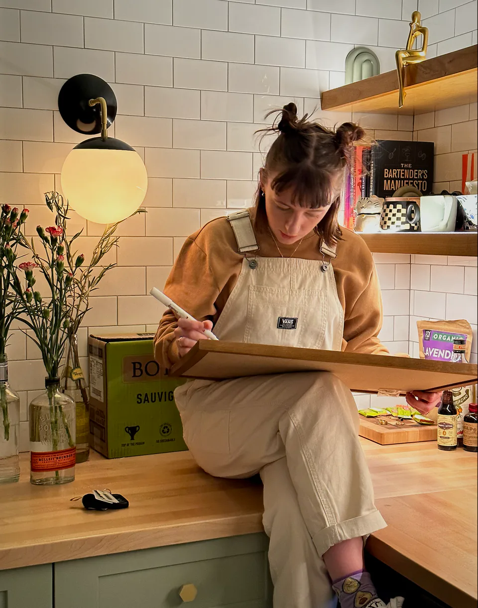 Sarah drawing in a sketchbook at a kitchen counter, tucked up on the bench under a round lamp.